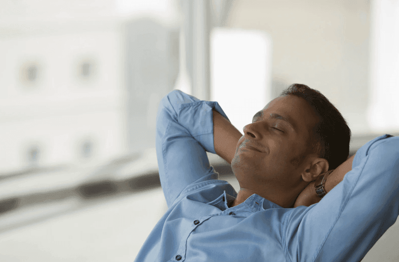 Person in blue shirt lounging with arms behind head, sitting by a large window.