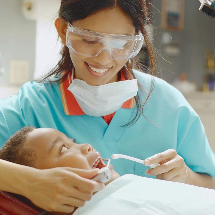 Dentist wearing protective glasses and a mask examining a young child's teeth with a dental tool.